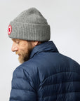 Man wearing a gray knit beanie with a Fjällräven logo and a blue puffer jacket on a light gray background