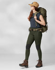 woman wearing blue top, green tights with reinforced knees and Fjällräven logo, a brown cap, carrying a trekking backpack