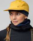 Person wearing a yellow cap and dark jacket with a visible Fjällräven logo on a gray background