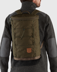 Person wearing a dark green backpack with a visible arctic fox logo on a light gray background