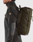 Person wearing a dark gray jacket with a green backpack on a light gray background