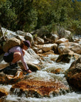 Person with a backpack collecting water from a stream in a forested area