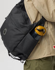 Person wearing a black backpack with a yellow handle, holding a beige jacket with a visible fjallraven logo.