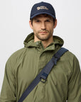 Man wearing a green raincoat and navy cap with a fjallraven logo on a light gray background