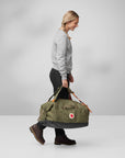 Person carrying a green duffel bag with a visible Fjällräven logo on a plain background