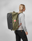 Person wearing a green and gray backpack with a visible Fjällräven logo on a plain background