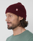 Man wearing a maroon beanie with a Fjällräven logo on a light gray background