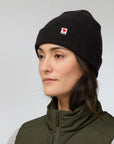 Woman wearing a black knit beanie with a Fjällräven logo on a light gray background