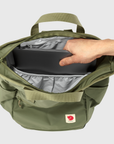Green backpack with a hand reaching inside to place a black item, on a light gray background
