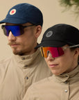 Two people wearing sunglasses and caps against a beige background