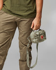 Person wearing green shirt and beige pants holding a camouflage bag with a red logo.