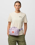 Person wearing a beige t-shirt and green pants with a purple waist bag on a light gray background