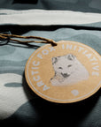 Round wooden tag with an arctic fox illustration and 'Arctic Fox Initiative' text on a camouflage fabric background.