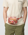 Person wearing a beige t-shirt and green pants with a red and brown patterned waist bag.
