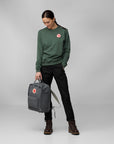 Person wearing a green sweatshirt and black pants, holding a gray backpack with a red logo on a gray background