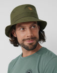 Man wearing a green bucket hat with a arctic fox logo on a plain background