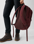 Person holding a maroon backpack with black pants and brown boots on a light gray background