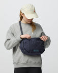 Person wearing a gray sweatshirt and beige cap holding a navy blue bag against a white background