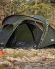 Green camping tent set up in a forest with cooking equipment nearby.