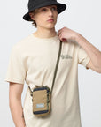 Person wearing a beige t-shirt with a logo and carrying a small bag on a plain background