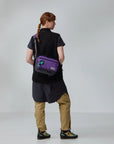 Person wearing a black shirt and beige pants with a purple bag on a white background