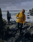 woman wearing yellow raincoat jacket with Fjällräven logo holding a camera and walking on stone surface