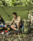Two people camping outdoors, cooking over a small fire under a green tarp.