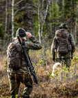 Two hunters with backpacks and rifles walking through a forest.