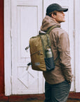 Man wearing a brown jacket and clay backpack with a red wooden door in the background