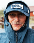 Person wearing a blue raincoat and cap with Fjällräven logo outdoors.