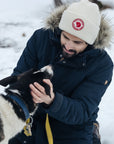 man wearing navy parka with fur hood and a white beanie playing with his dog