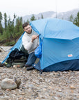 Man setting up a blue tent in a natural outdoor setting with mountains and trees in the background.