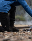 Person in blue jacket and black pants cooking over a campfire by a body of water.
