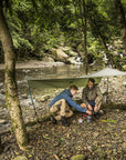 Two people setting up a tent in a forest by a stream