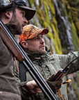 Two hunters in camouflage gear with rifles and communication devices in a forest setting.