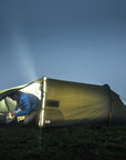 Person inside a tent with a flashlight and another person outside on a grassy hill at night.