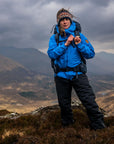 Person in blue jacket and black pants standing on a mountain with a backpack, surrounded by misty mountains.