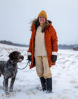 Woman in an orange coat and yellow beanie standing in a snowy field with a dog.