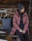 Person wearing a pink coat sitting in front of a wooden cabin with a blue bag beside them.
