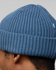 Blue knit beanie with a small flag emblem worn by a person on a light gray background