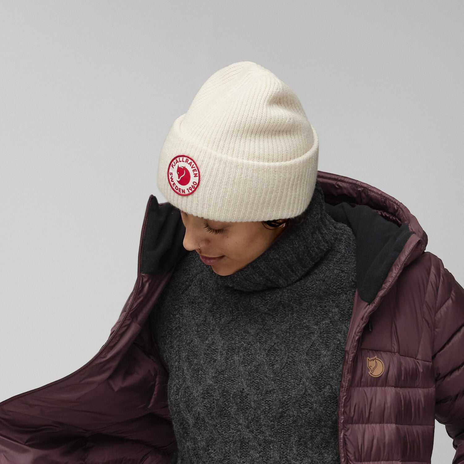 Person wearing a white beanie with a Fjällräven logo and a maroon puffer jacket on a gray background