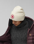 Person wearing a white beanie with a Fjällräven logo and a maroon puffer jacket on a gray background