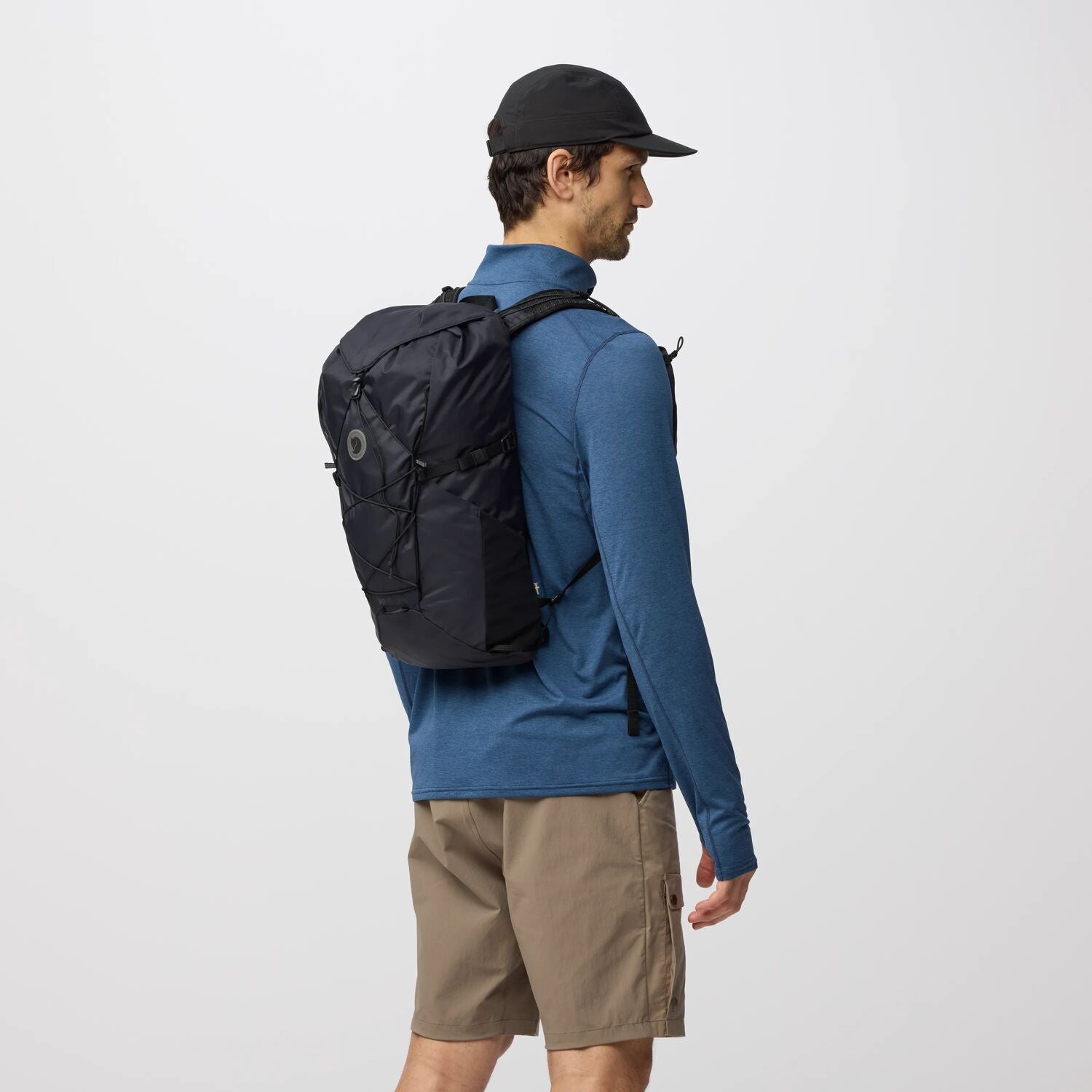 Man wearing a black backpack, blue shirt, and beige shorts on a white background
