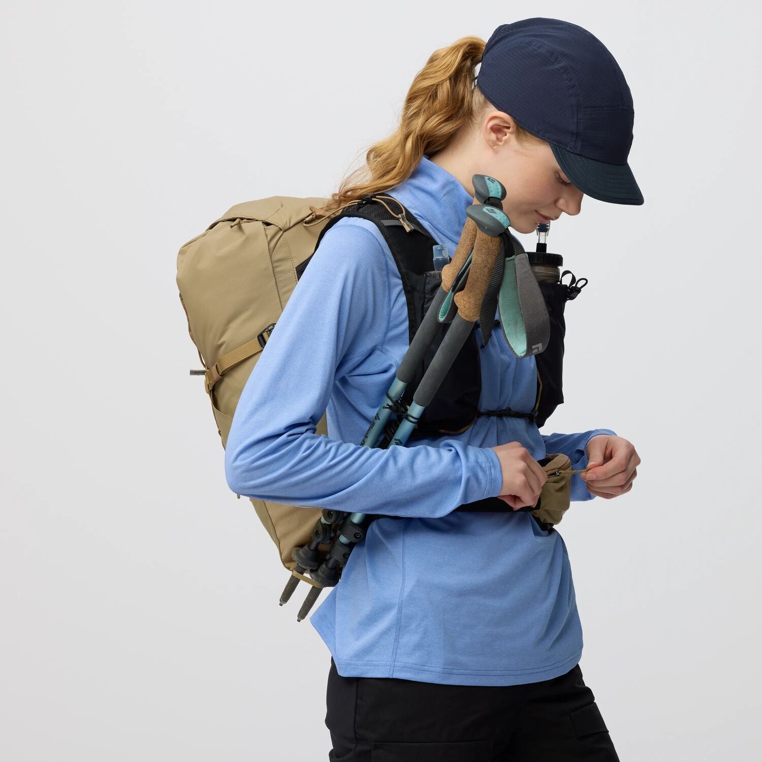 Person wearing a blue shirt, black cap, and carrying hiking gear on a plain background