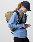 Person wearing a blue shirt, black cap, and carrying hiking gear on a plain background