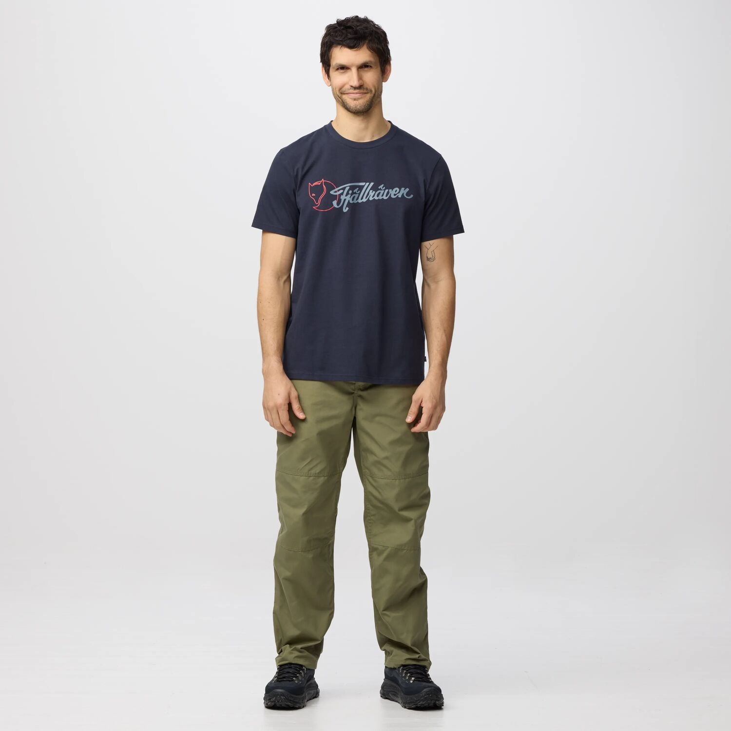 Man wearing a navy t-shirt with a Fjällräven  logo and green pants on a white background