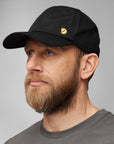 Man wearing a black cap with a Fjällräven logo on a gray background