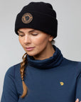 Woman wearing a black knit beanie with a Fjällräven logo and a blue turtleneck sweater with a Fjällräven logo on a gray background