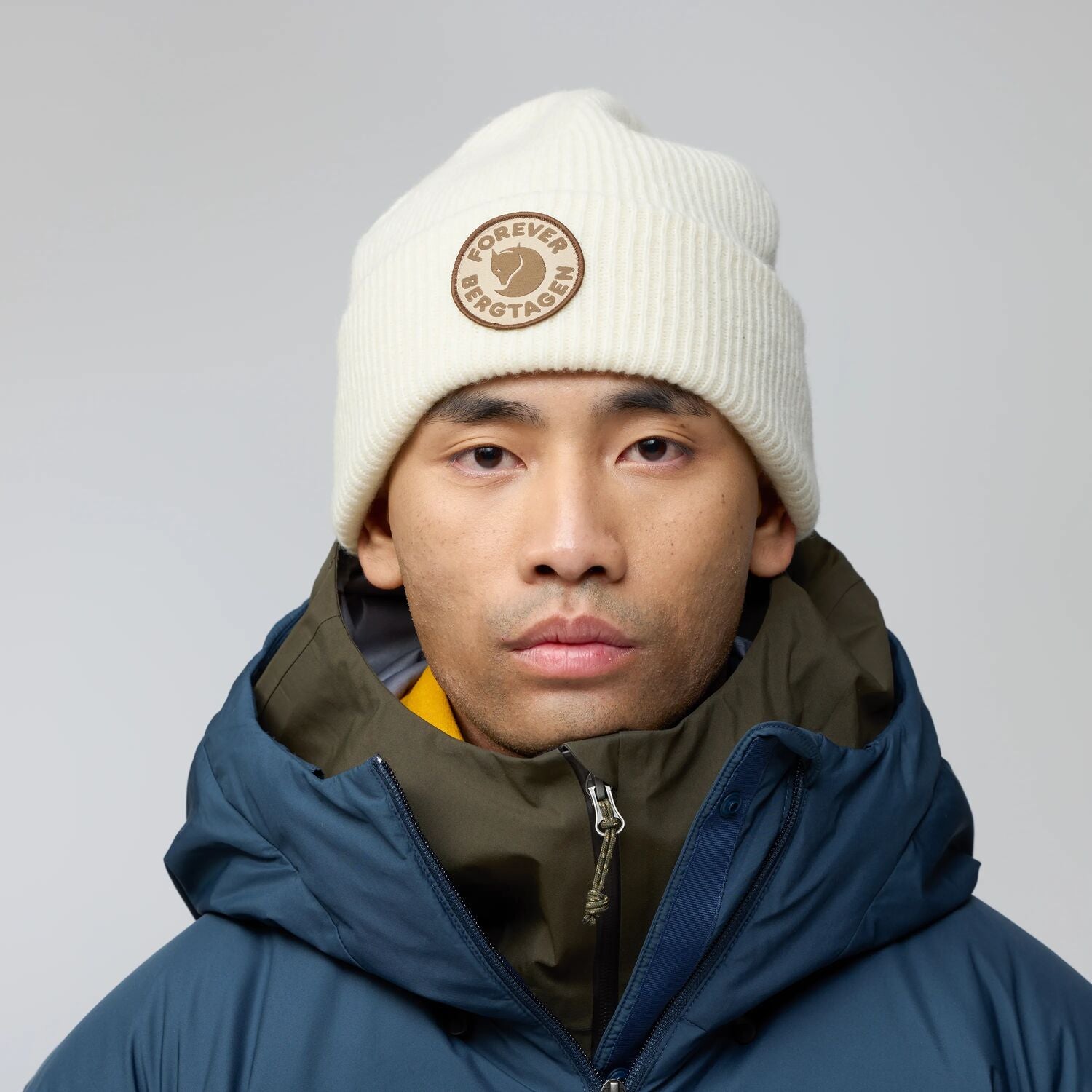 Person wearing a white beanie with a Fjällräven logo and a blue jacket on a gray background