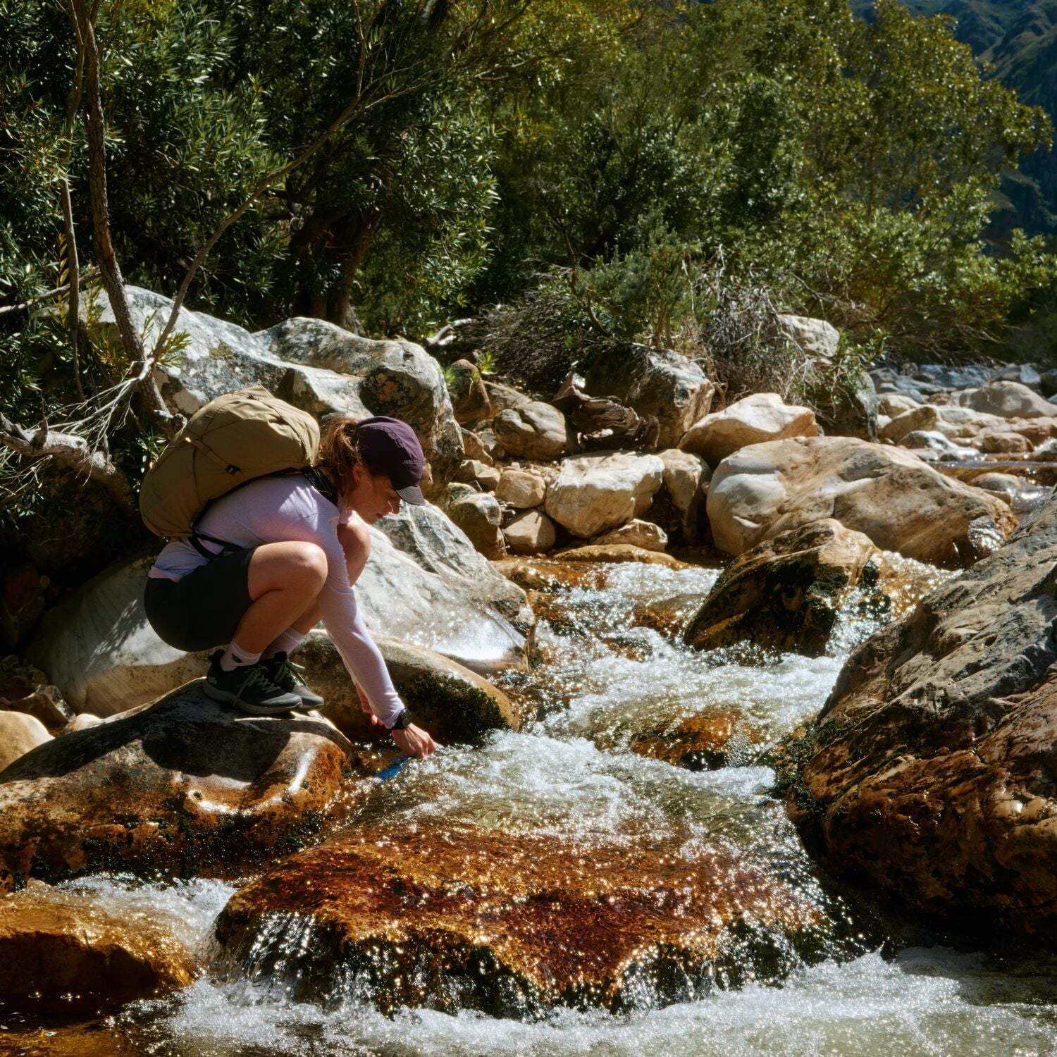 Person with a backpack collecting water from a stream in a forested area